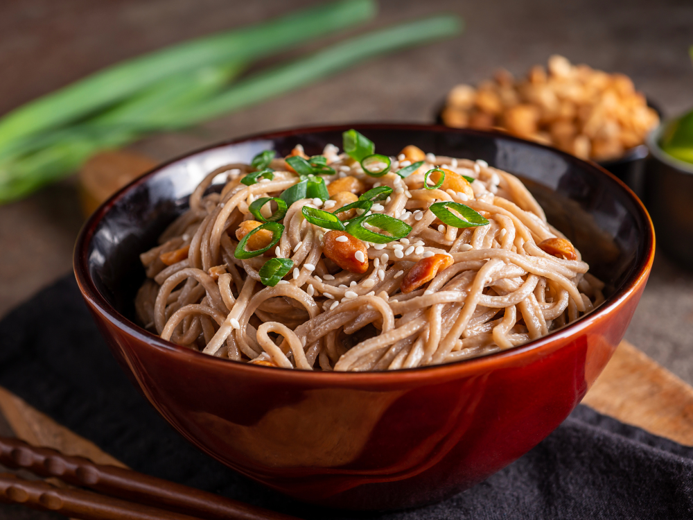 A bowl filled with sesame peanut noodles topped with sliced green onions, chopped peanuts, and sesame seeds. 