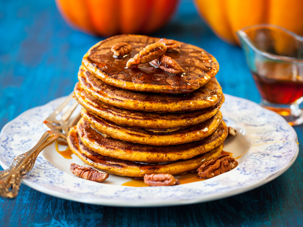 Stack of fluffy pumpkin pancakes topped with butter and syrup, served on a plate on a blue, wooden table. The pancakes are golden brown and have visible pieces of pumpkin and spices. A fork is placed next to the plate.