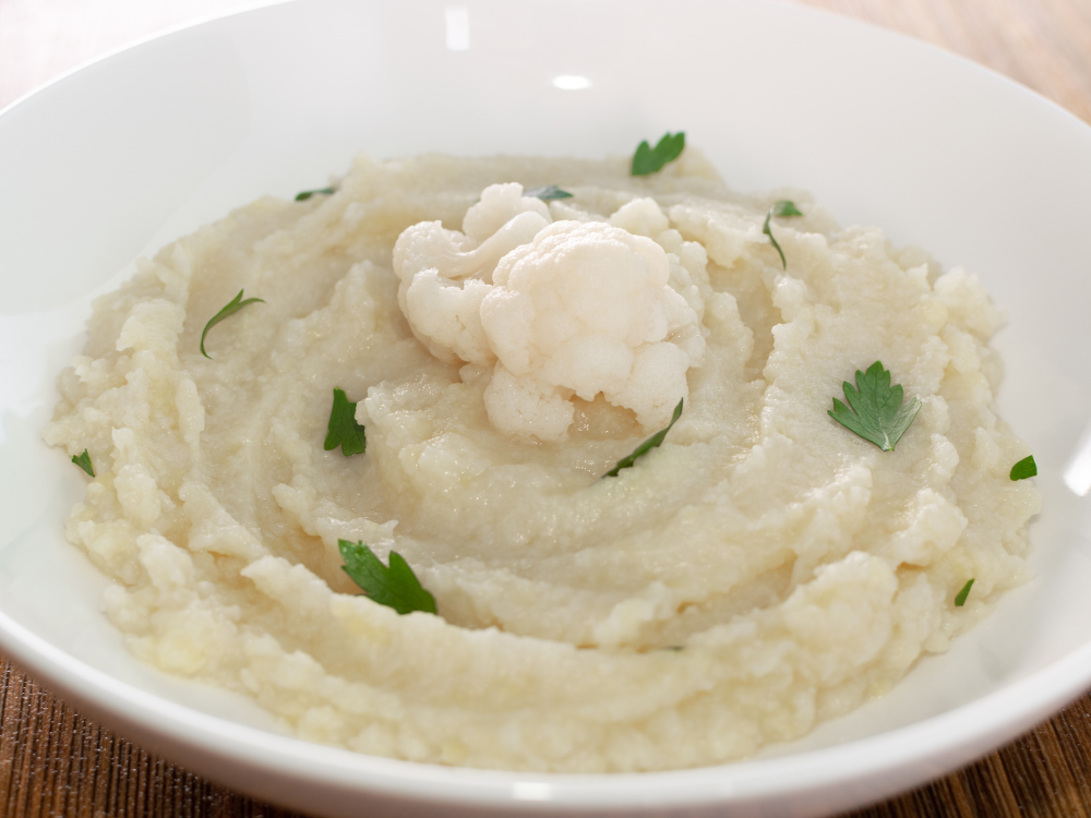 A bowl of creamy white mashed potatoes with small pieces of cauliflower throughout. The dish is topped with a sprinkle of chopped green herbs and a few small cloves of roasted garlic.