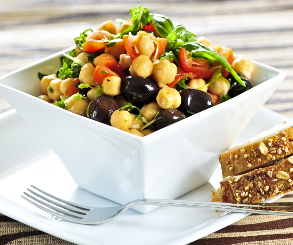 Image of a colorful sweet bean salad made with garbanzo beans, fresh spinach, chopped dates, crumbled feta cheese, and other delicious ingredients. The salad is served in a white bowl and garnished with chopped herbs.