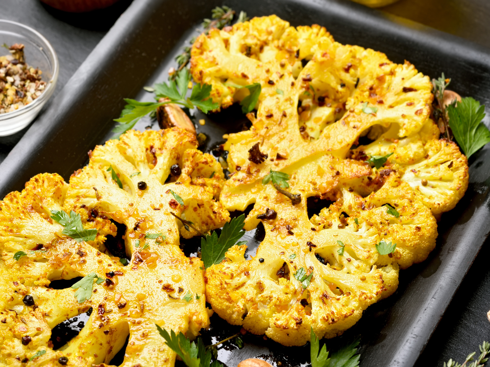 A close-up photo of golden-brown cauliflower florets arranged on a baking sheet. The cauliflower is coated in a crispy breadcrumb and herb mixture and baked until tender on the inside.