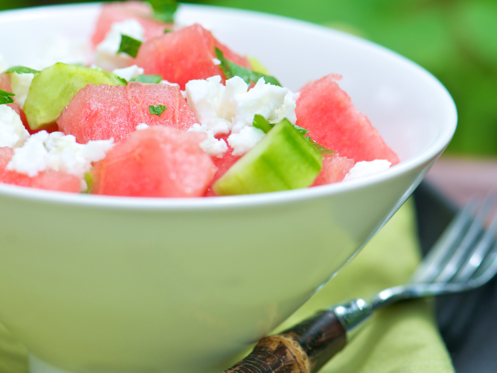 The image shows a fresh and colorful salad composed of sliced cucumbers, diced watermelon, crumbled feta cheese, and chopped mint leaves.