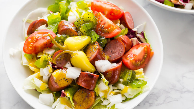A close-up photo of a colorful salad in a bowl, featuring diced tomatoes, sliced pickles, chopped onions, sweet relish, and yellow mustard, all tossed with pieces of grilled hot dog and mixed greens.